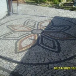 Okrągły wzór kwiatowy ułożony z kostki granitowej w odcieniach szarości, brązu i czerni, otoczony szarą kostką, widoczny cień rzucany przez budynek.